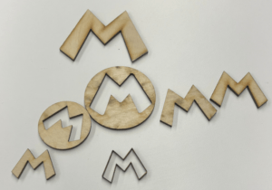 Laser cut pieces of the Mario “M” logo of varying sizes, some as outlines of the “M”, some as just the “M”, and some as circles with the “M” cut out of the middle laid out on a white background. 